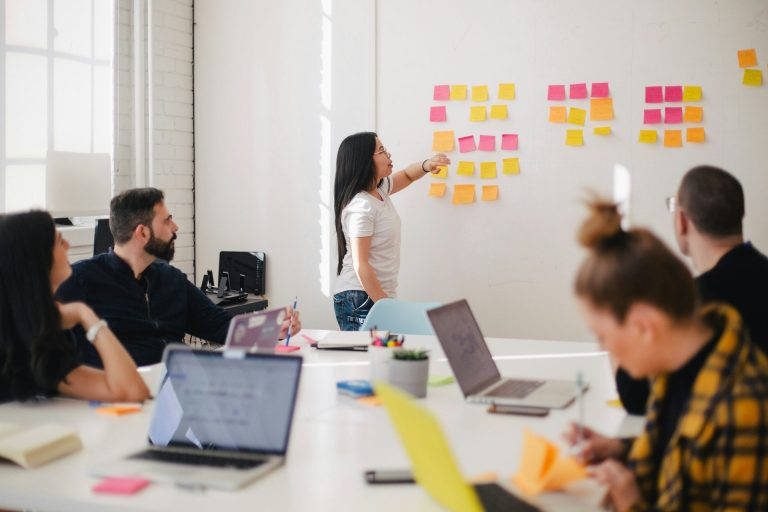 Eine Gruppe von Menschen sitzt in einer Schulung an einem Tisch während eine Person Notizen auf einem Whiteboard präsentiert.